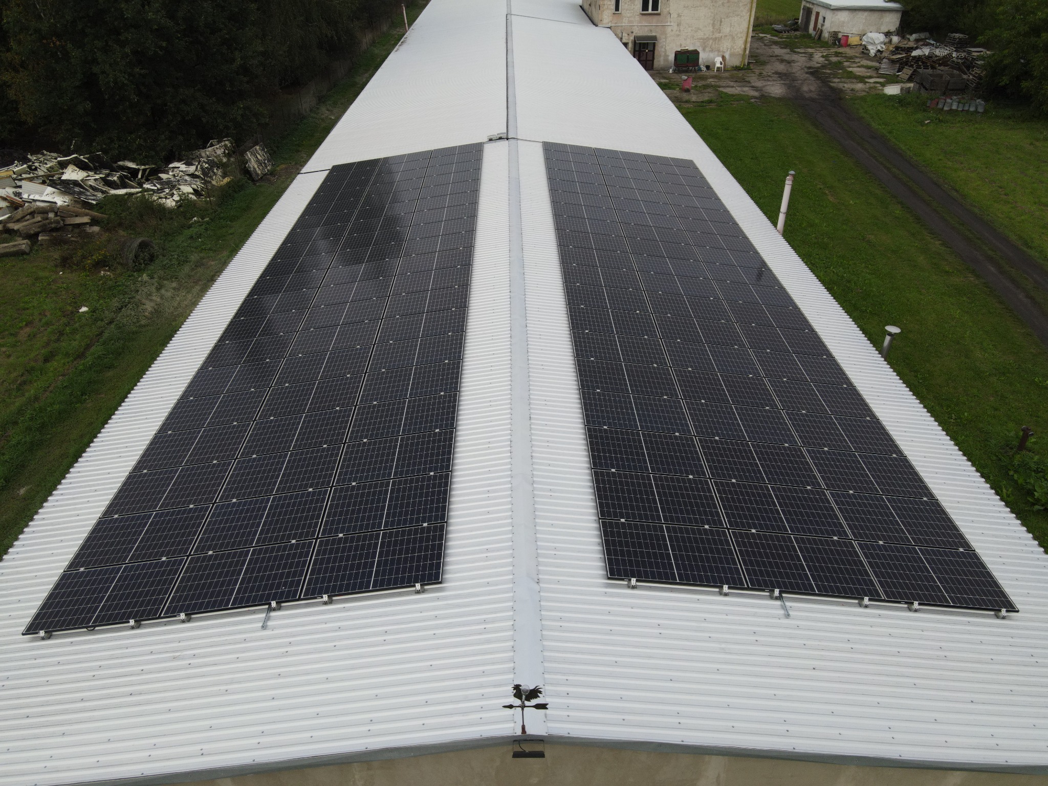 Widok z góry na dwa rzędy paneli słonecznych na falistym, metalowym dachu budynku gospodarczego; na dachu wiatrowskaz w kształcie koguta, w tle zieleń i zabudowania.