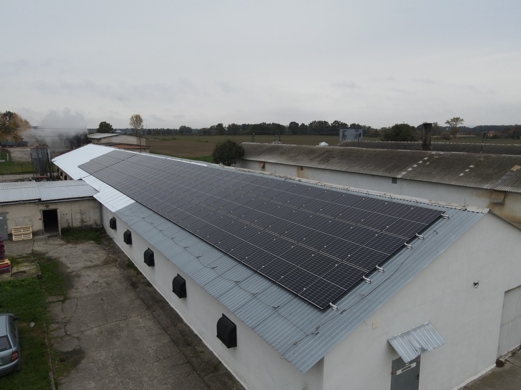 Instalacja fotowoltaiczna na dachu budynku gospodarczego z blachy falistej; widok z góry na panele słoneczne w układzie prostokątnym, szary samochód osobowy i palety w pobliżu budynku, oraz komin...