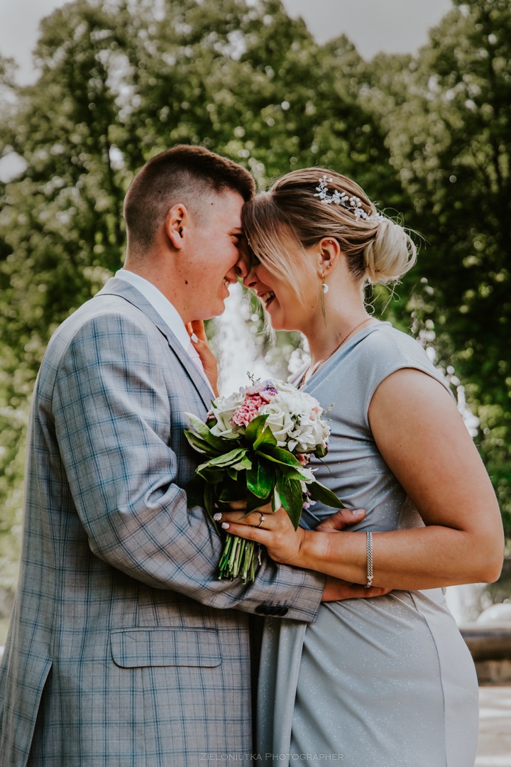 Uśmiechnięta para, panna młoda z bukietem i ozdobą we włosach, w objęciach na tle zieleni.