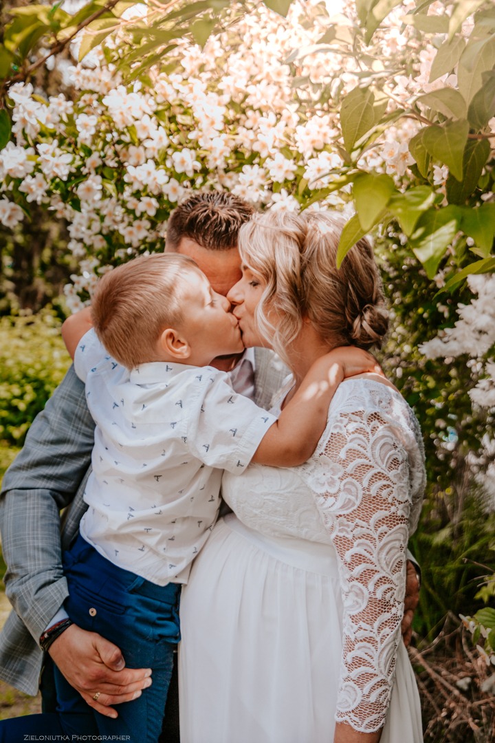 Ciepłe ujęcie rodziny: ojciec w szarej marynarce trzyma syna w niebieskich spodniach i białej koszuli, obaj całują ciężarną matkę w białej koronkowej sukience na tle kwitnących białych kwiatów.