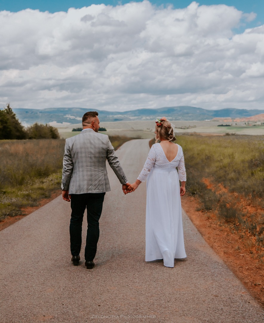 Para młoda, trzymając się za ręce, idzie drogą w Kudowie-Zdroju; panna młoda w białej sukni z koronkowymi rękawami i kwiatami we włosach, pan młody w szarej marynarce w kratę, widok z tyłu, w tle...