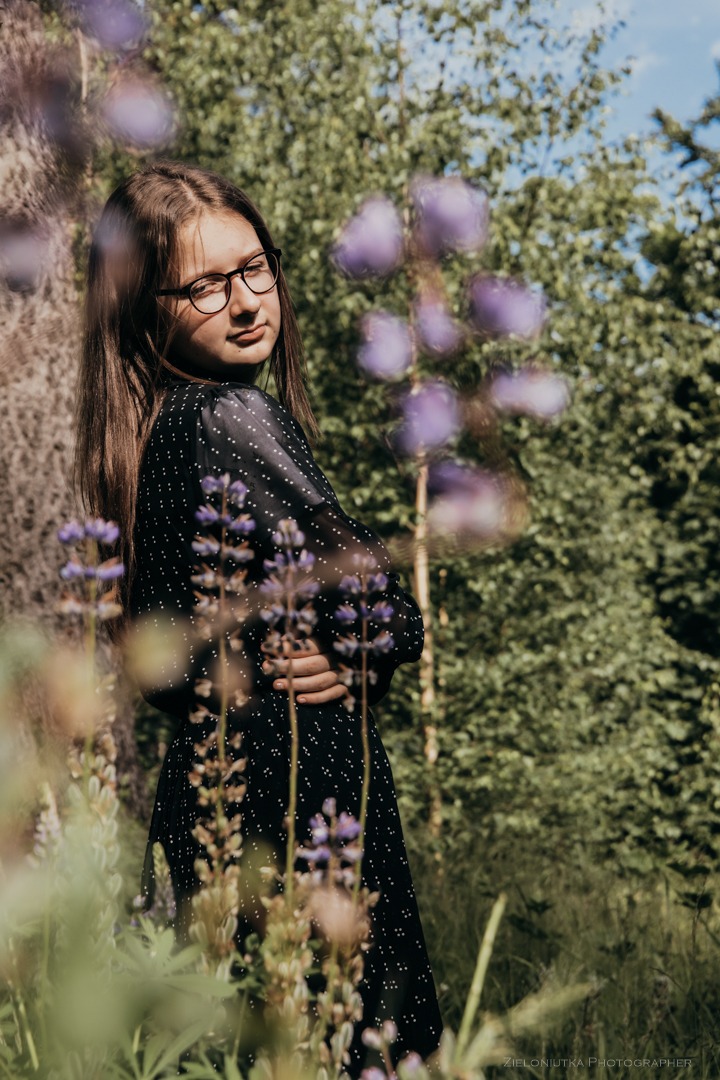 Portret nastolatki w okularach, pozującej w kwitnącym łubinie na tle drzew, w czarnej sukience w białe kropki.