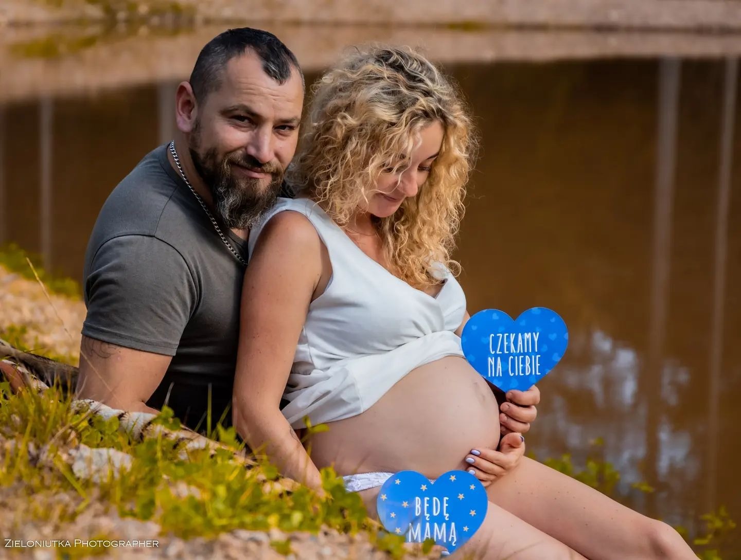 Przyszła mama z partnerem pozują na tle jeziora, trzymając niebieskie serca z napisami 'Czekamy na Ciebie' i 'Będę Mamą'.