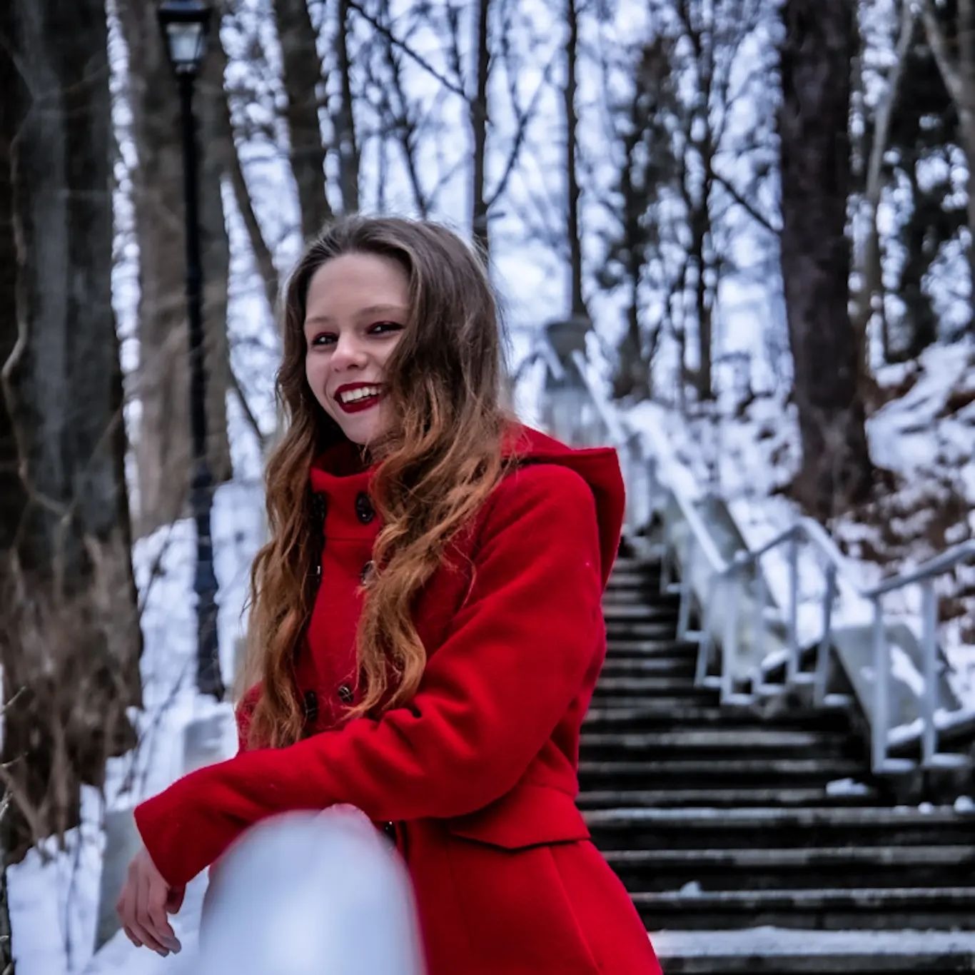 Uśmiechnięta młoda kobieta w czerwonym płaszczu z kapturem, oparta o metalową barierkę na tle zimowego parku ze schodami.
