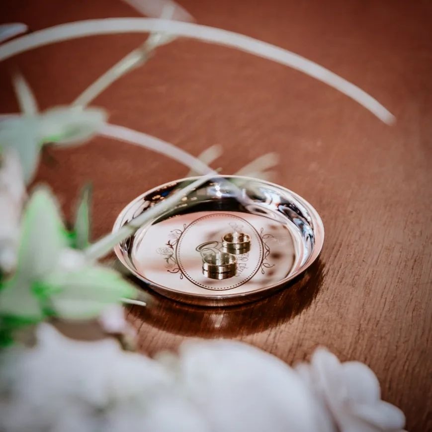 Dwa złote obrączki na srebrnej tacy z ozdobnym grawerem, leżące na drewnianym stole, z delikatnymi kwiatami w tle i na pierwszym planie.