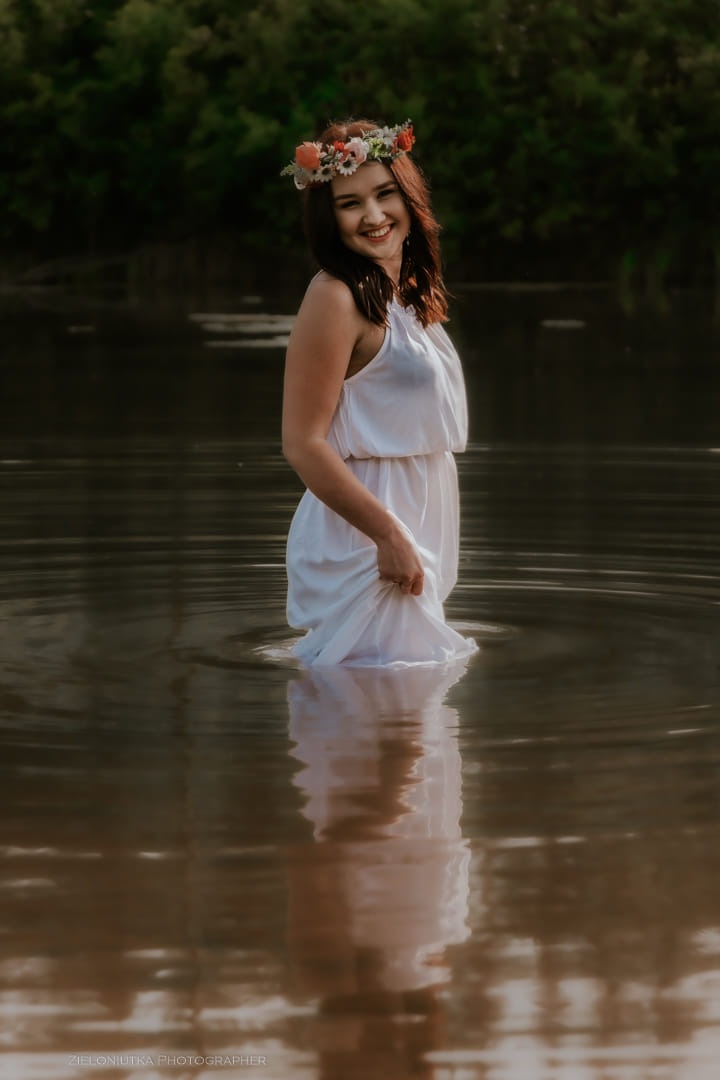 Uśmiechnięta kobieta w białej sukience i wianku na głowie, stojąca w wodzie po kolana, z widocznym odbiciem w tafli wody.