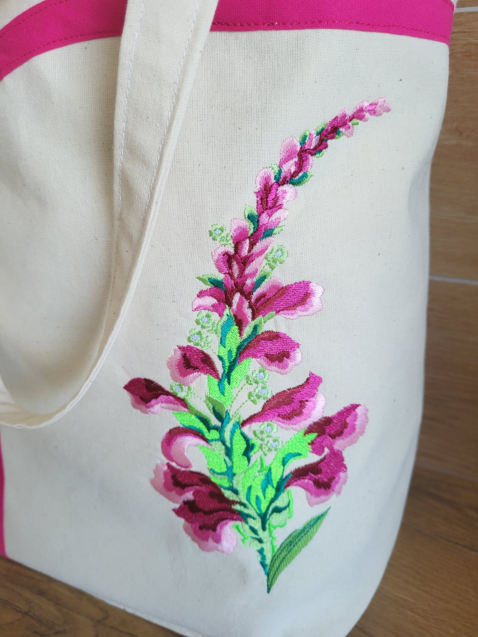 Ecru torba bawełniana z haftowanym motywem kwiatowym w odcieniach różu i zieleni, wykończona różową lamówką na górze.