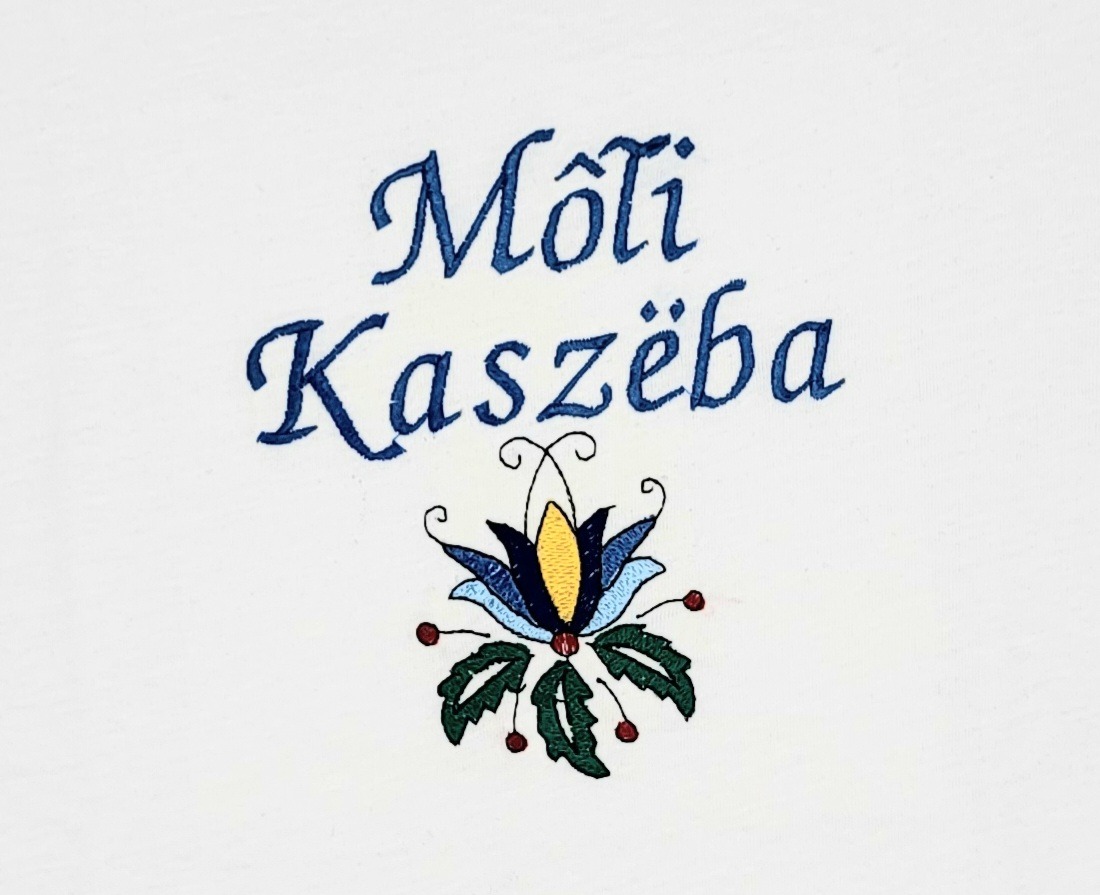 Biała tkanina z niebieskim haftem przedstawiającym napis 'Môti Kaszëba' oraz stylizowany kwiat w kolorach niebieskim, żółtym, zielonym i czerwonym.
