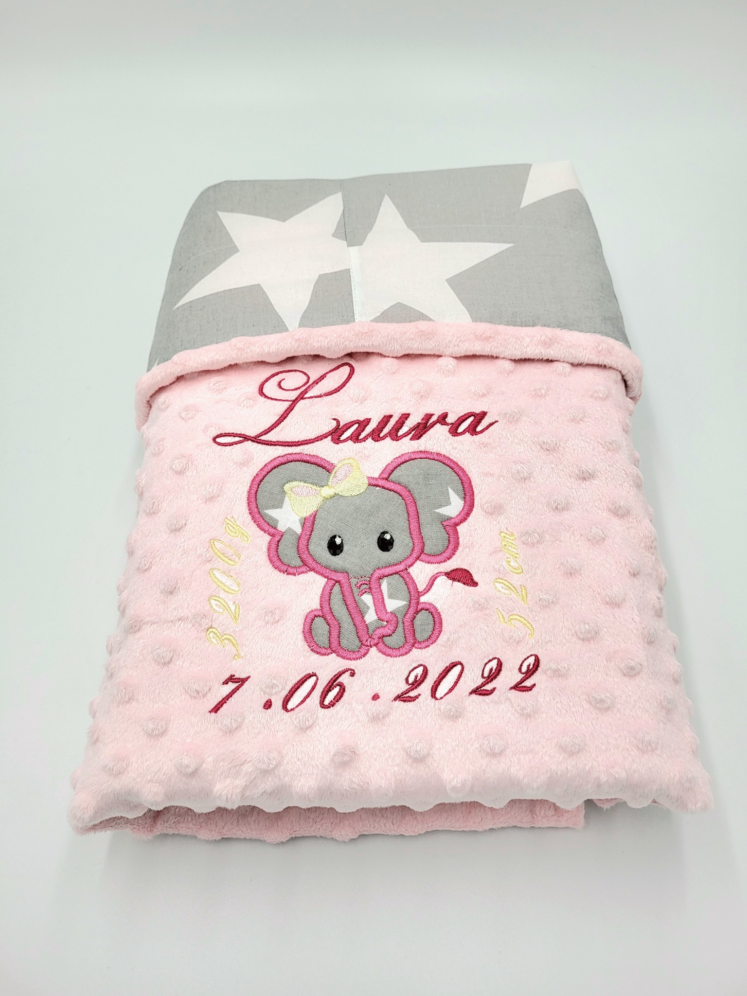 Różowy kocyk z wypukłą fakturą, haftowanym imieniem Laura, słoniem z kokardą i datą 7.06.2022, leżący na białym tle, z widoczną szarą podszewką w białe gwiazdy.