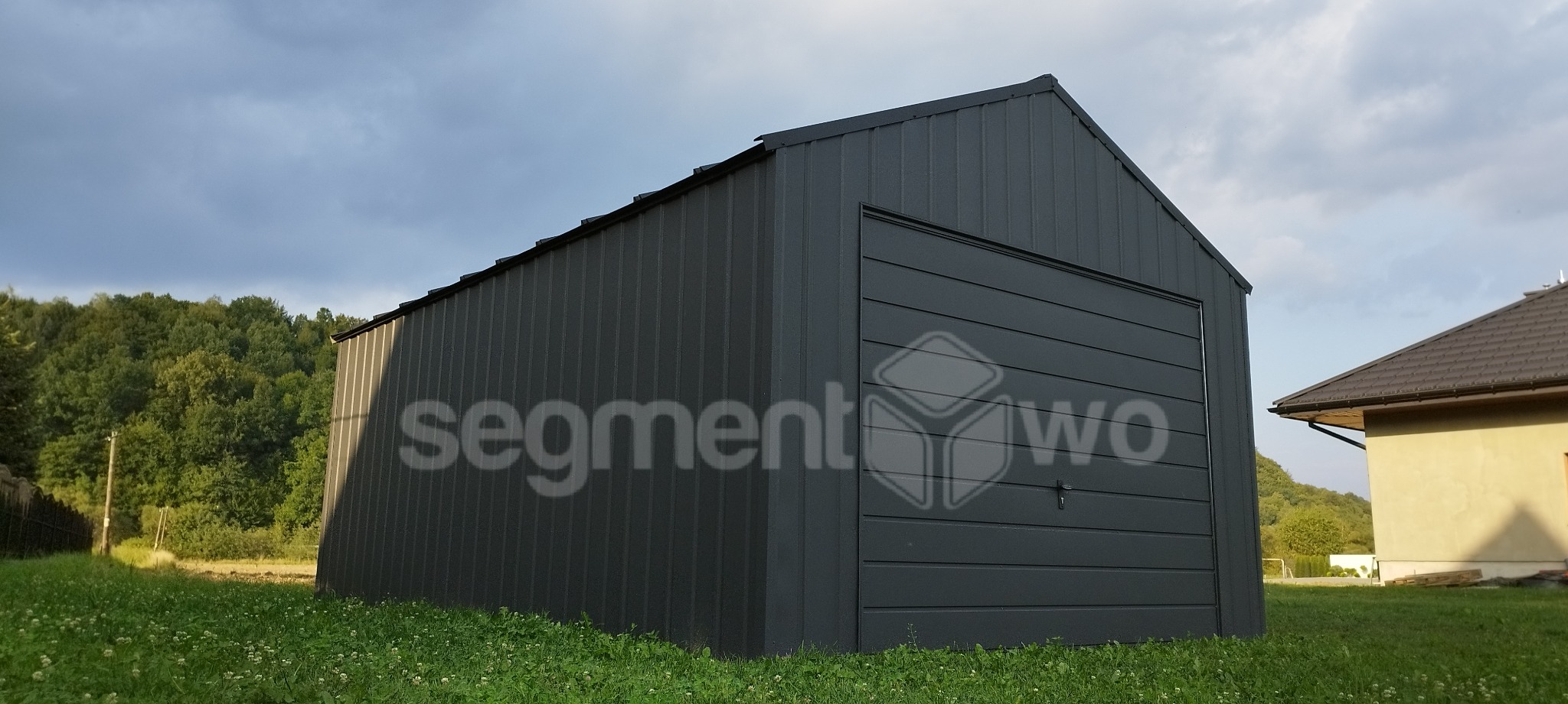 Ciemnoszary garaż z blachy trapezowej z bramą segmentową, widoczny z niskiej perspektywy na tle zieleni i pochmurnego nieba, z nałożonym półprzezroczystym logo firmy na elewacji.