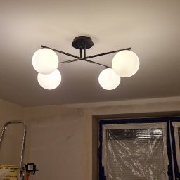 Krok po kroku - Czarny, nowoczesny żyrandol z czterema białymi, kulistymi kloszami zamontowany na suficie w trakcie remontu; okno zabezpieczone folią malarską, drabina malarska w rogu pokoju.