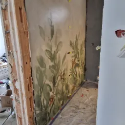 Ściana w trakcie remontu z dekoracyjnym malowaniem roślinnym, zabezpieczona folią podłoga, widoczne żółte kliny poziomujące i fragment surowego muru.