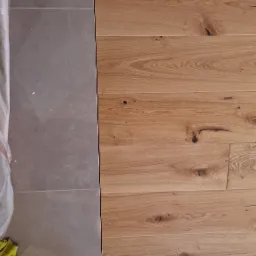 Połączenie płytek podłogowych imitujących kamień i drewnianej podłogi z widocznymi sękami, zabezpieczone klinami przy ścianie.