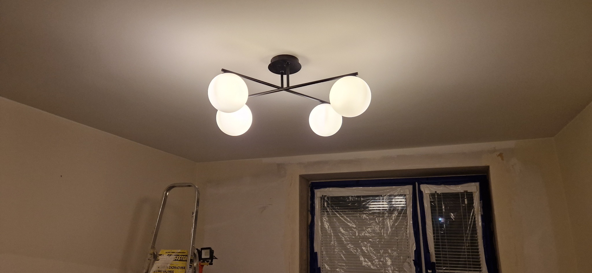 Czarny, nowoczesny żyrandol z czterema białymi, kulistymi kloszami zamontowany na suficie w trakcie remontu; okno zabezpieczone folią malarską, drabina malarska w rogu pokoju.