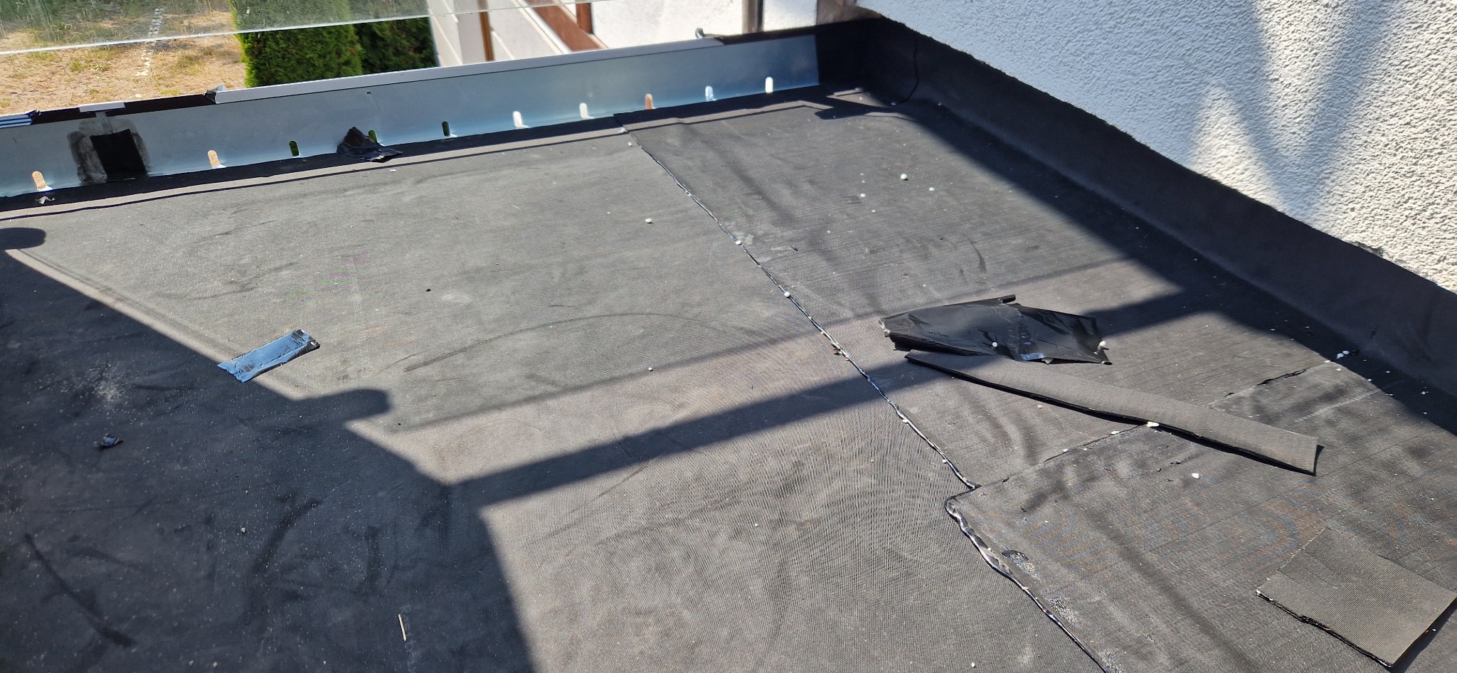 Uszkodzona papa na płaskim dachu z widocznymi łatami i fragmentami oderwanego materiału, metalowa listwa okapowa w tle.