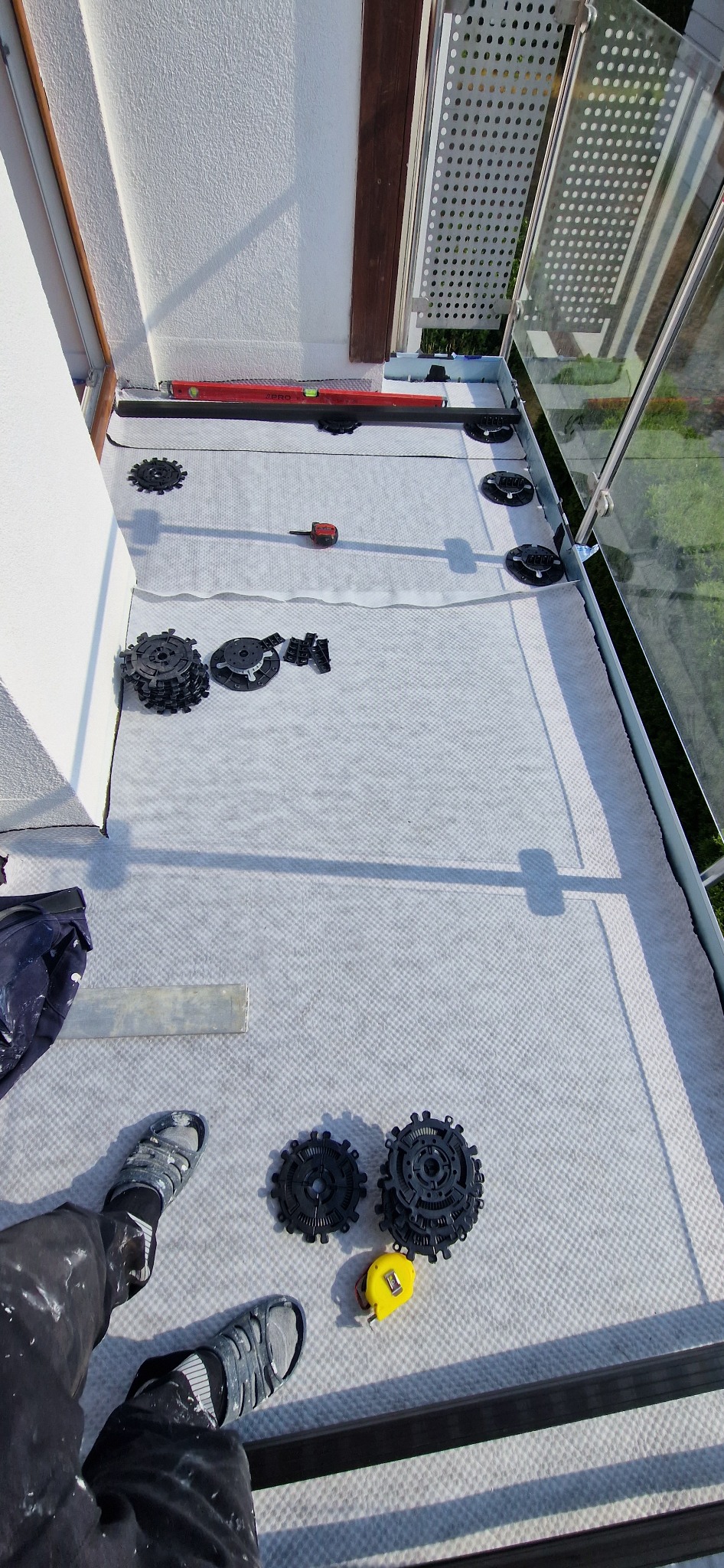 Biała mata izolacyjna na balkonie z rozłożonymi regulowanymi wspornikami tarasowymi, poziomica, miarka i robocze ubranie, przygotowanie do montażu podłogi.