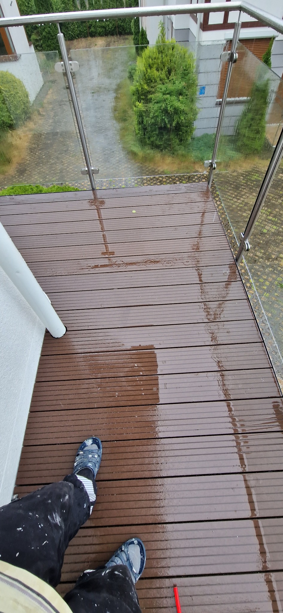Mokry taras z desek kompozytowych po deszczu, widoczne stopy osoby w ubrudzonych farbą spodniach i klapkach, szklana balustrada z metalowymi słupkami, w tle zieleń i budynek.
