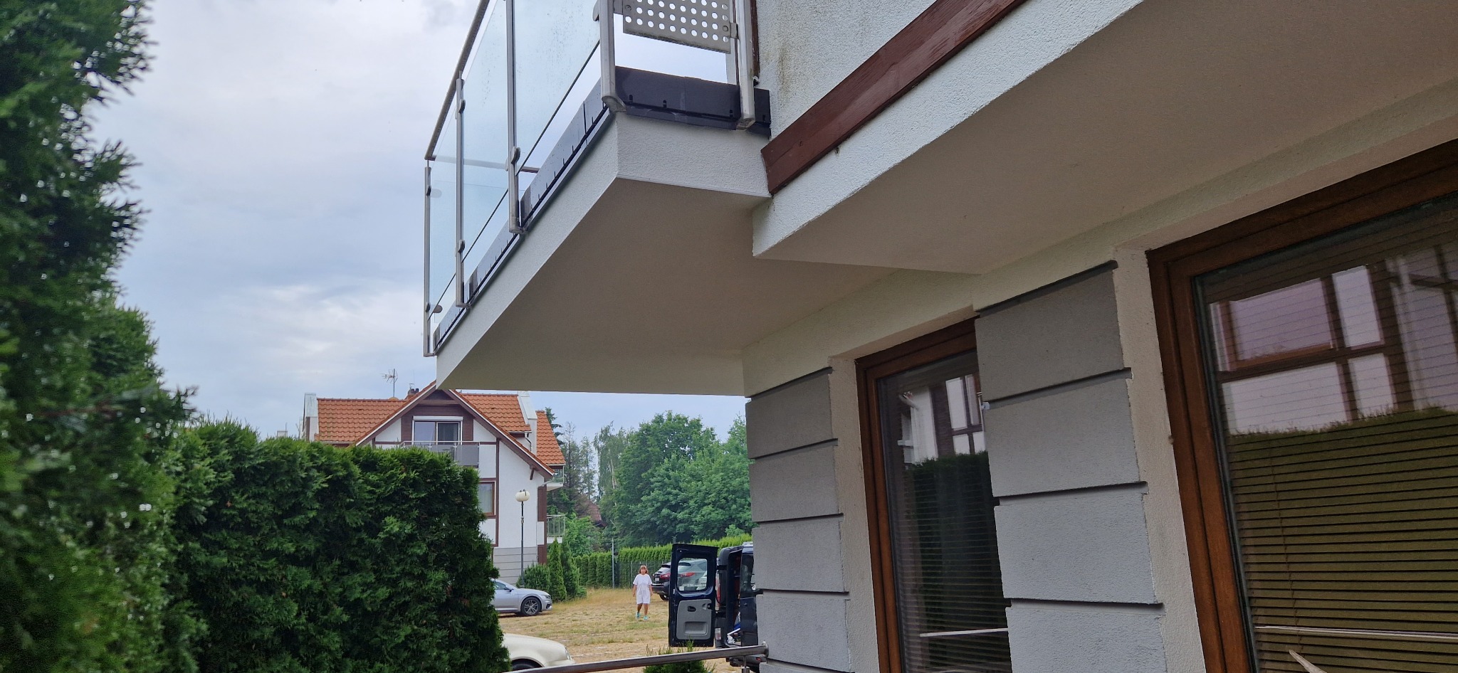 Elewacja budynku po odświeżeniu koloru, z widocznym balkonem z przeszkloną balustradą, oknem w brązowej ramie i fragmentem zielonego żywopłotu w tle.