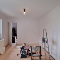 RAFTI-Rafał Szymczak - Białe ściany po malowaniu w pokoju z oknem, grzejnikiem i drabiną. Widoczne narzędzia malarskie i otwarte drzwi do innego pomieszczenia. Jasne wnętrze po odświeżeniu.
