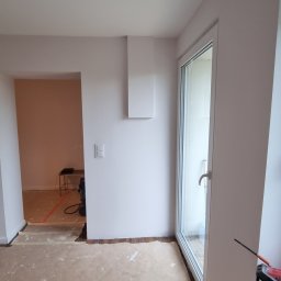 RAFTI-Rafał Szymczak - Wnętrze po malowaniu ścian na jasny odcień, widoczne zabezpieczone podłogi, okno PCV i grzejnik. Na ścianie zamontowany biały element dekoracyjny.