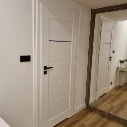 Białe drzwi wewnętrzne z czarną klamką, widoczne również w odbiciu lustrzanym, na tle paneli podłogowych.