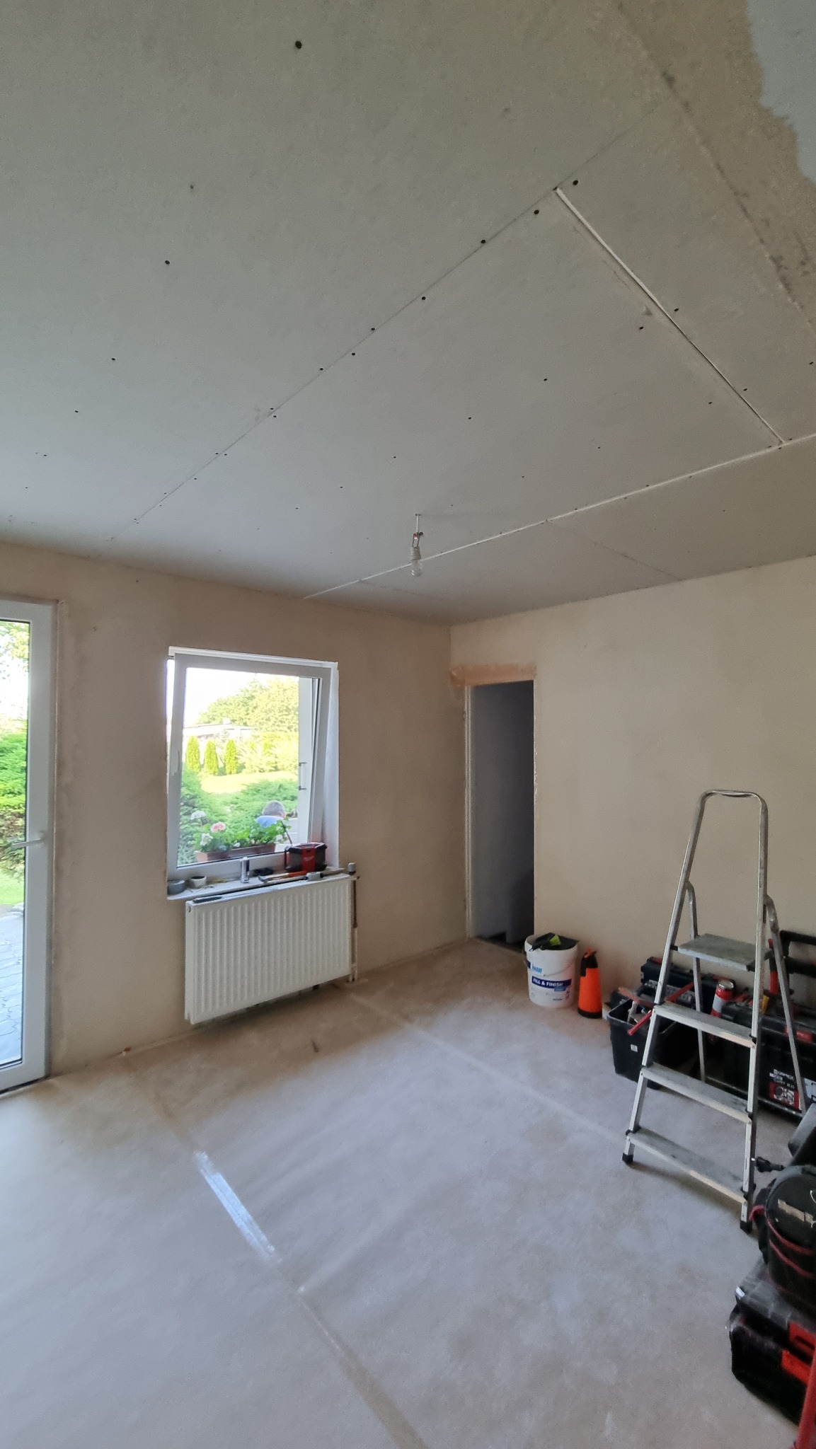 Pokój w trakcie remontu: ściany przygotowane pod malowanie, sufit z płyt karton-gips, drabina i narzędzia malarskie. Widok na okno z zielenią na zewnątrz.