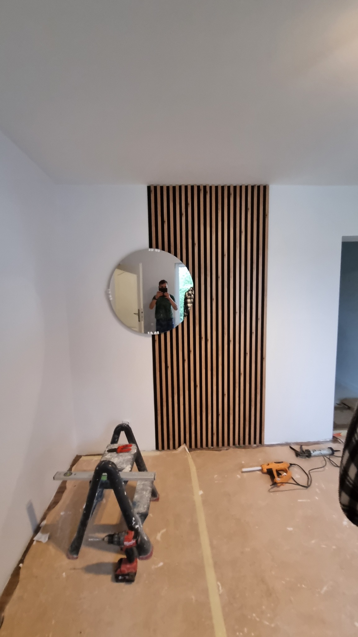 Montaż dekoracyjnych paneli ściennych z drewna. Widoczne narzędzia, drabina i odbicie osoby w okrągłym lustrze na ścianie. Wnętrze w trakcie wykończenia.