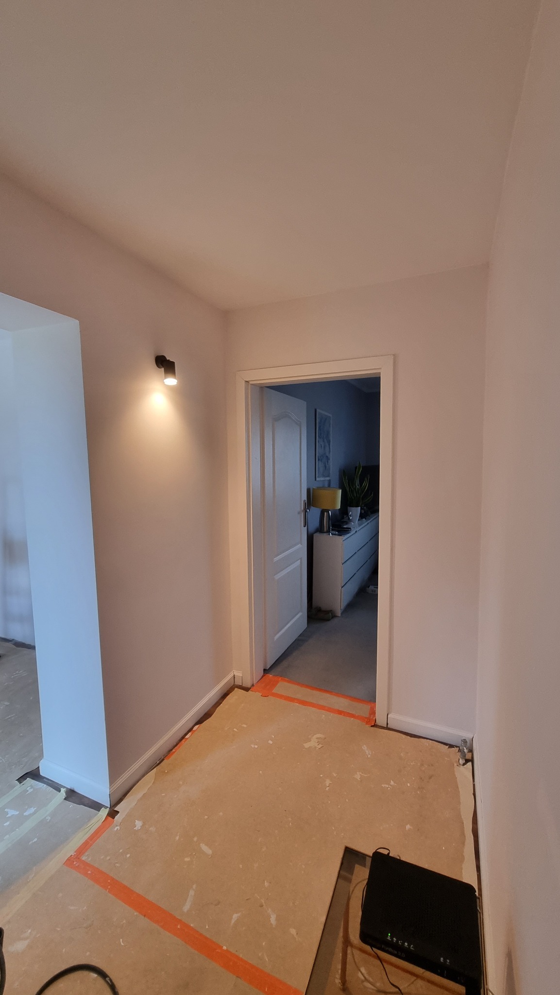 Pokój w trakcie remontu: świeżo pomalowane ściany w odcieniach bieli, zabezpieczona podłoga, widoczne oświetlenie punktowe i otwarte drzwi do kolejnego pomieszczenia.