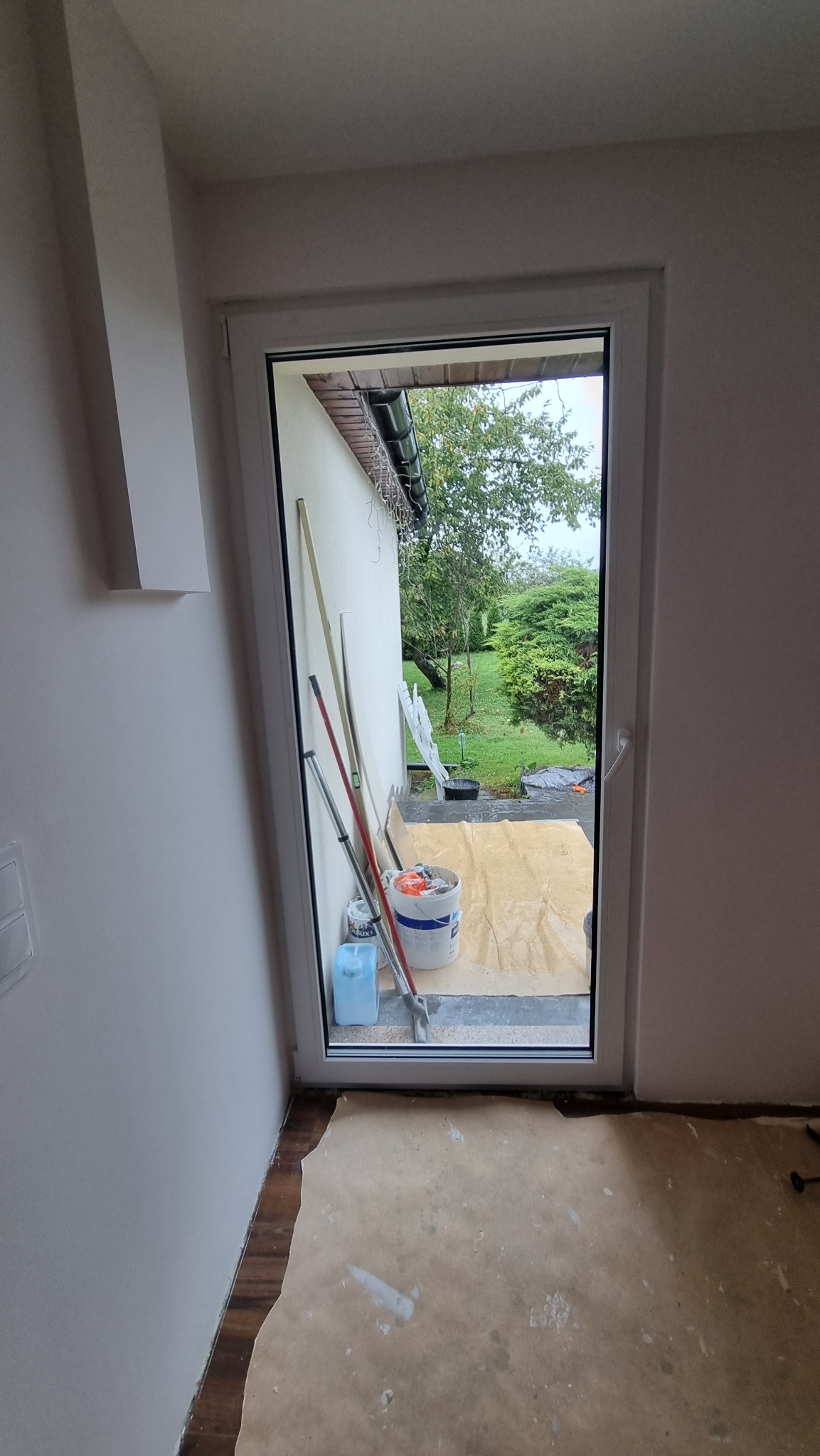 Białe okno PCV z widokiem na ogród, zabezpieczone folią i narzędziami malarskimi w trakcie prac wykończeniowych. Widoczne wiadro z farbą i wałki malarskie.