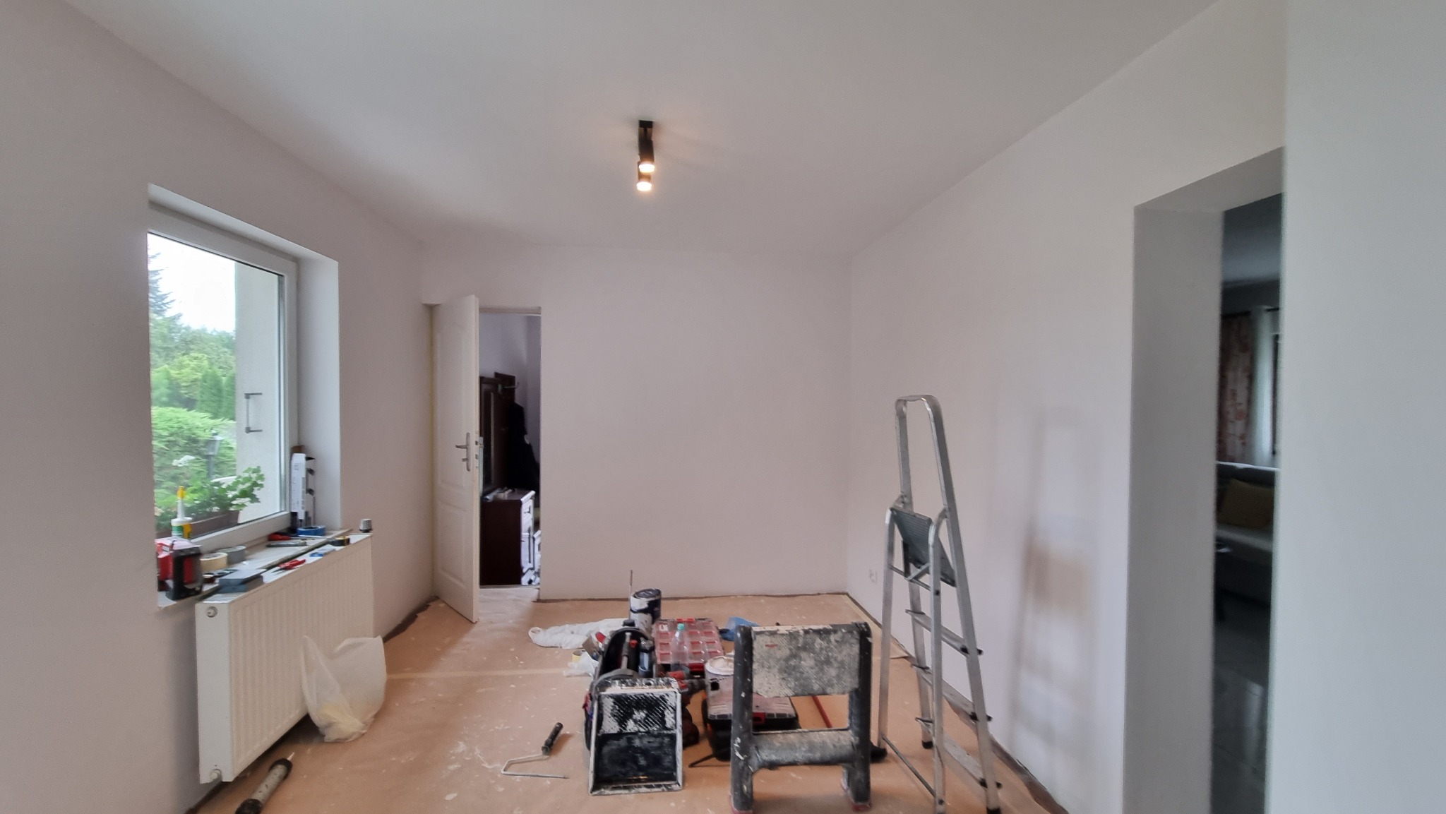 Białe ściany po malowaniu w pokoju z oknem, grzejnikiem i drabiną. Widoczne narzędzia malarskie i otwarte drzwi do innego pomieszczenia. Jasne wnętrze po odświeżeniu.