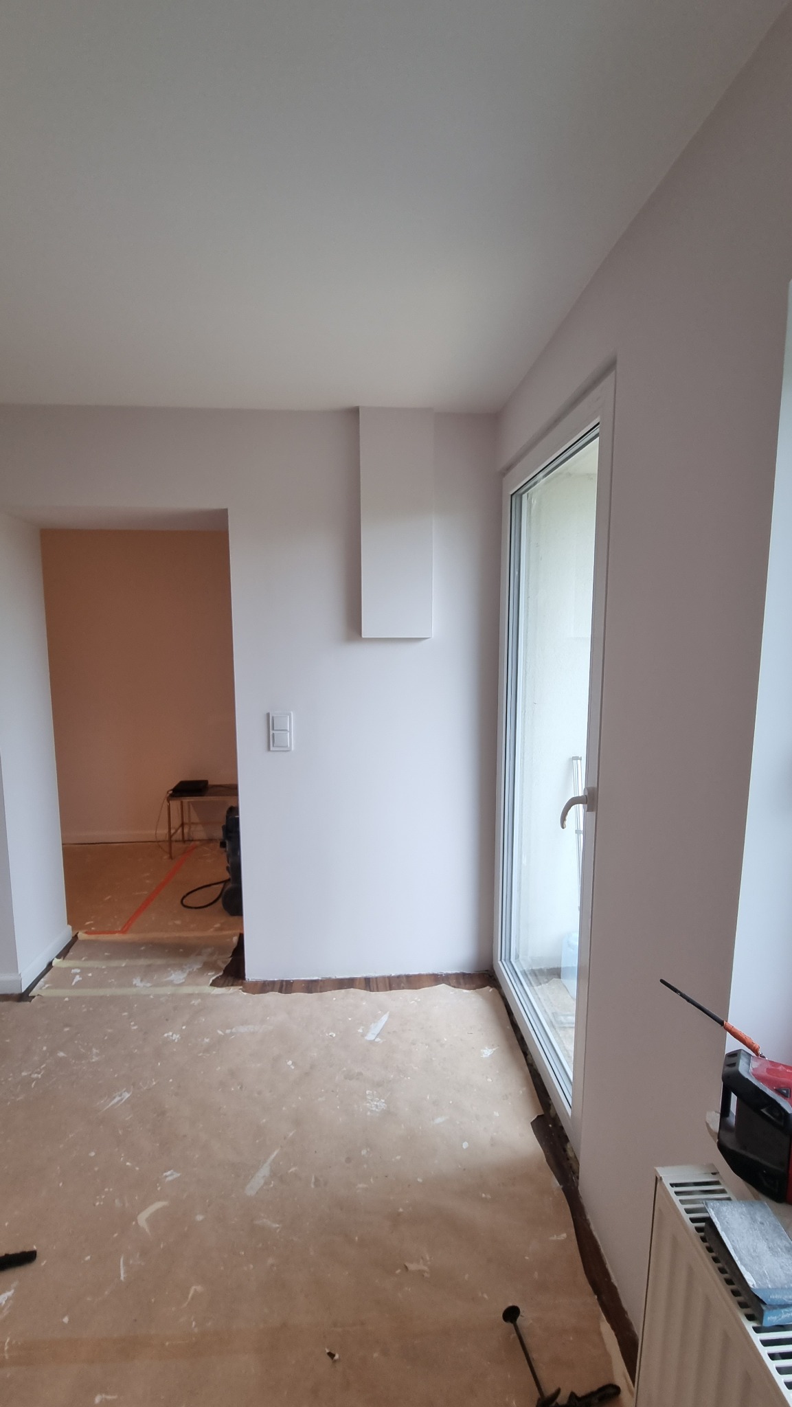 Wnętrze po malowaniu ścian na jasny odcień, widoczne zabezpieczone podłogi, okno PCV i grzejnik. Na ścianie zamontowany biały element dekoracyjny.