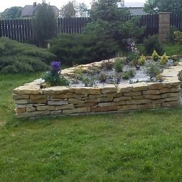 Visual Garden - Kaskadowa rabata kwiatowa z kamienia piaskowca w ogrodzie, z niskimi roślinami iglastymi i kwitnącymi bylinami, na tle trawnika i drewnianego płotu.