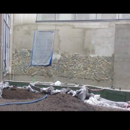 Visual Garden - Przygotowanie do założenia ogrodu zimowego w Lubinie: widoczna ściana wyłożona kamieniem, ziemia przygotowana pod nasadzenia, okno zabezpieczone folią malarską.