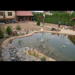 Visual Garden - Oczko wodne z kamienną kaskadą i roślinnością wodną w ogrodzie, z widocznym trawnikiem, drzewami i pracownikami wykonującymi prace wykończeniowe.