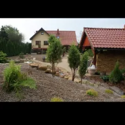 Nowo założony ogród z podwyższonymi rabatami, kamieniami ozdobnymi, młodymi drzewkami iglastymi i oczkiem wodnym w tle