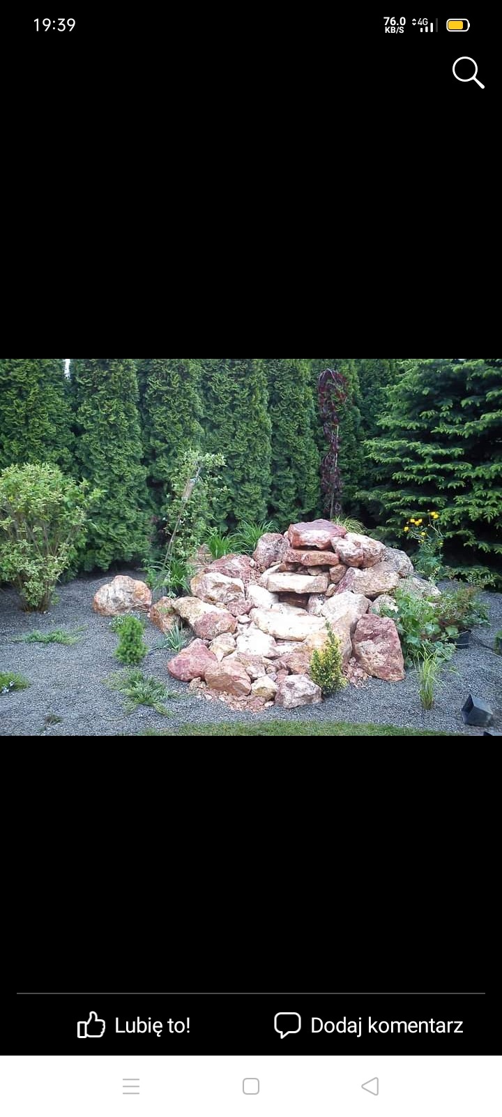 Ogród z kaskadą z kamieni, otoczoną iglakami i krzewami ozdobnymi, na podłożu z drobnego żwiru.