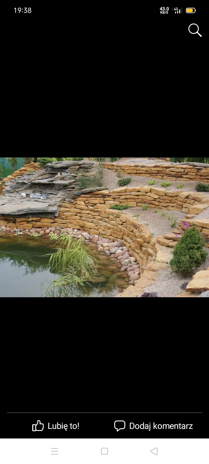 Oczko wodne z kaskadą z kamienia piaskowca, otoczone roślinnością, w tym trawami wodnymi i niskimi krzewami.