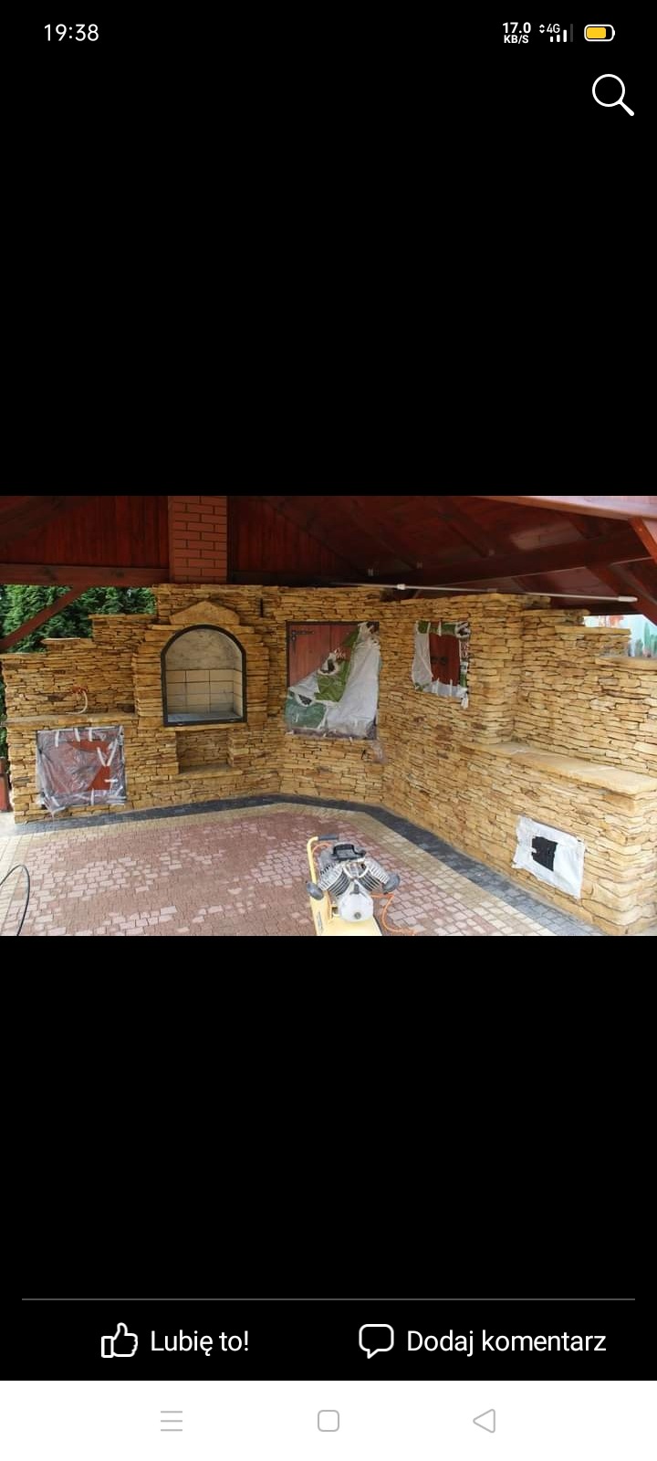 Budowa kamiennego grilla zewnętrznego w trakcie realizacji, z widoczną maszyną do zagęszczania podłoża i częściowo zakrytymi elementami konstrukcyjnymi.