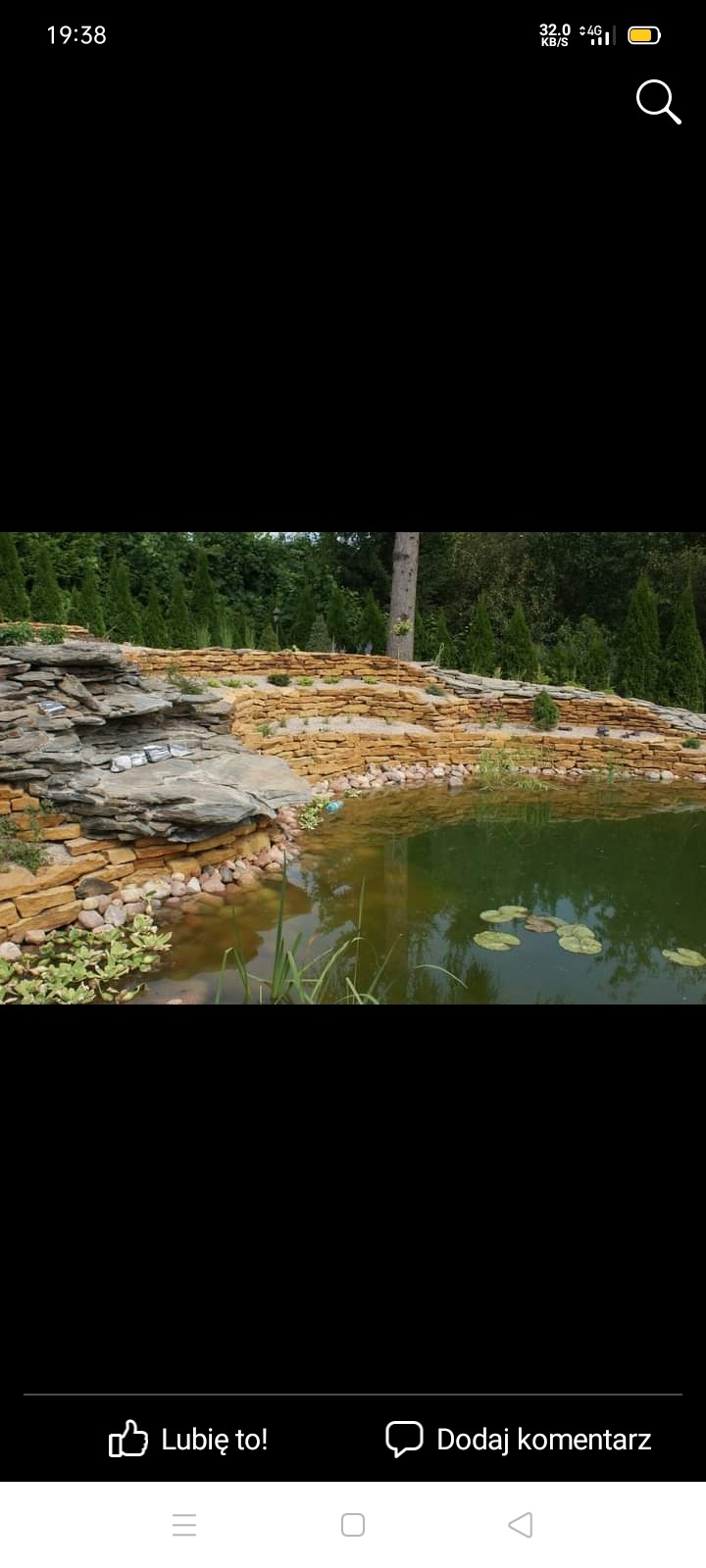 Oczko wodne z kamienną kaskadą i obudową z piaskowca, otoczone zielenią i drzewami iglastymi.