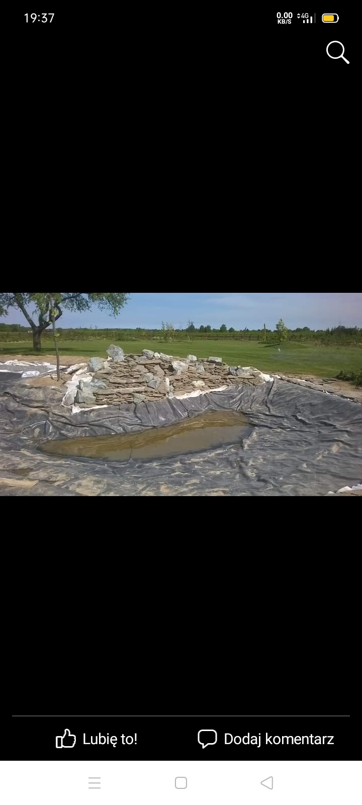 Budowa oczka wodnego w ogrodzie, widoczne ułożone kamienie, czarna folia i częściowo wypełniona woda, w tle trawnik i drzewa.