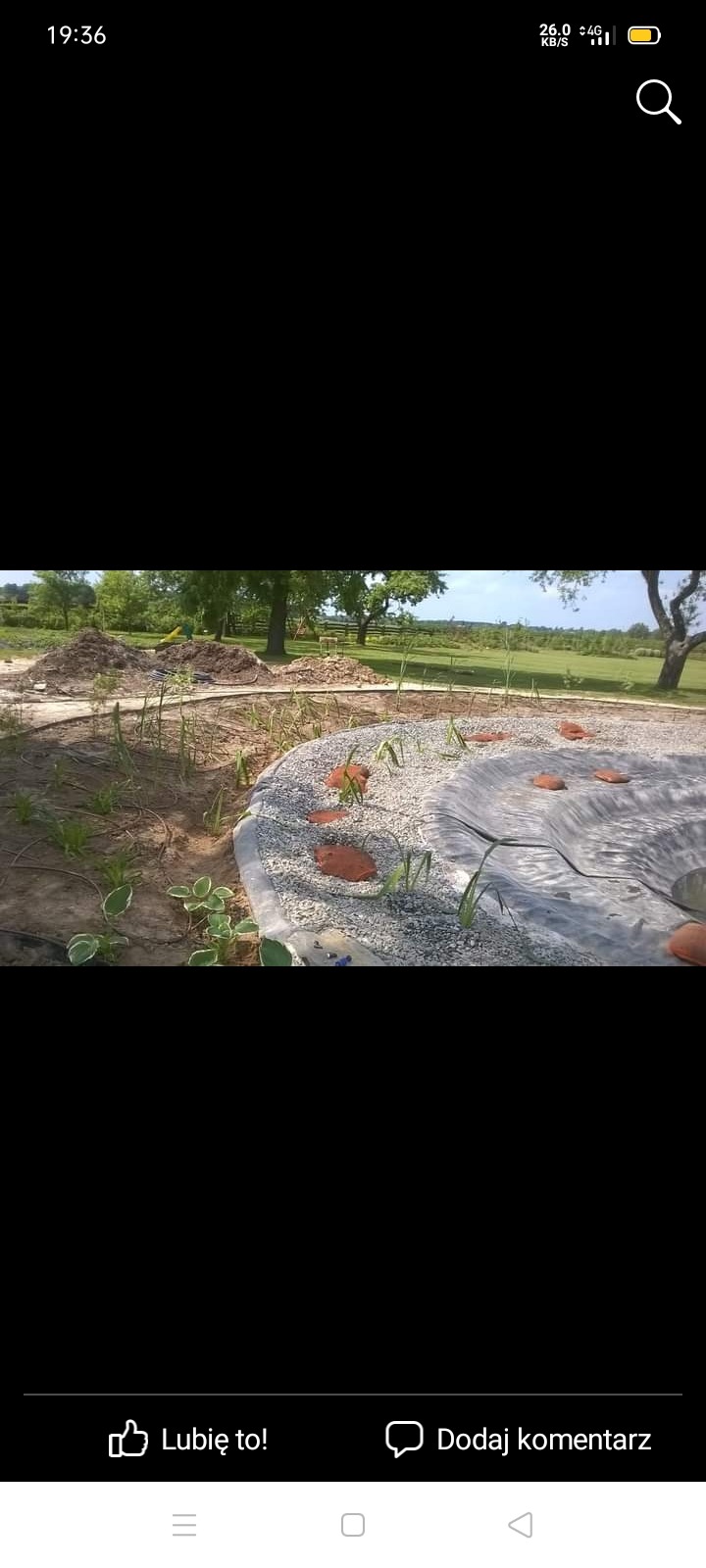 Budowa oczka wodnego z wyłożoną folią, obsypanego kamieniami i z elementami dekoracyjnymi z cegły, w tle trawnik i drzewa.