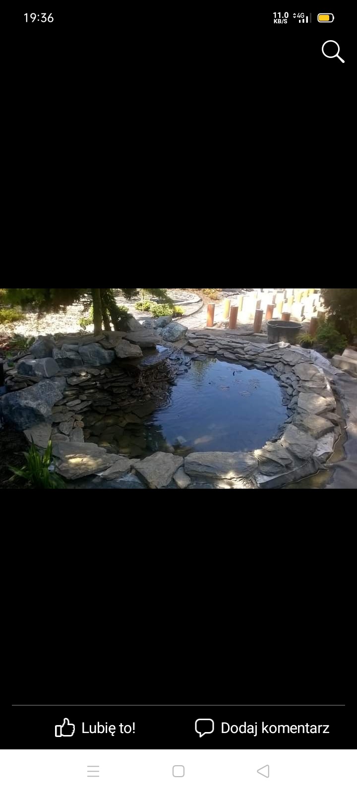 Okrągły, kamienny oczko wodne w ogrodzie, otoczone różnej wielkości szarymi kamieniami, z widocznymi elementami instalacji wokół.