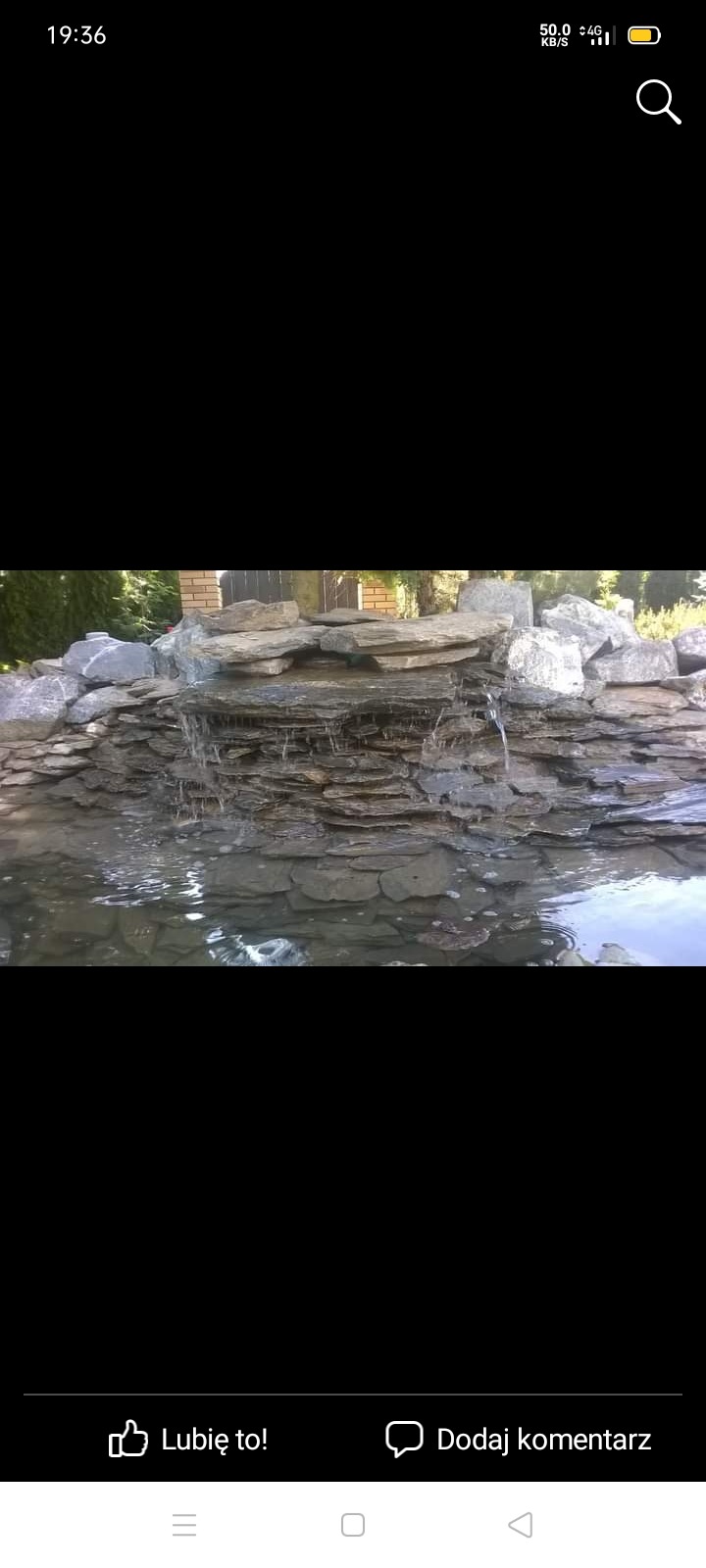 Kaskada wodna z kamienia łupkowego w ogrodzie, delikatny strumień wody spływa po warstwach skalnych do oczka wodnego, z ceglanym murem w tle.