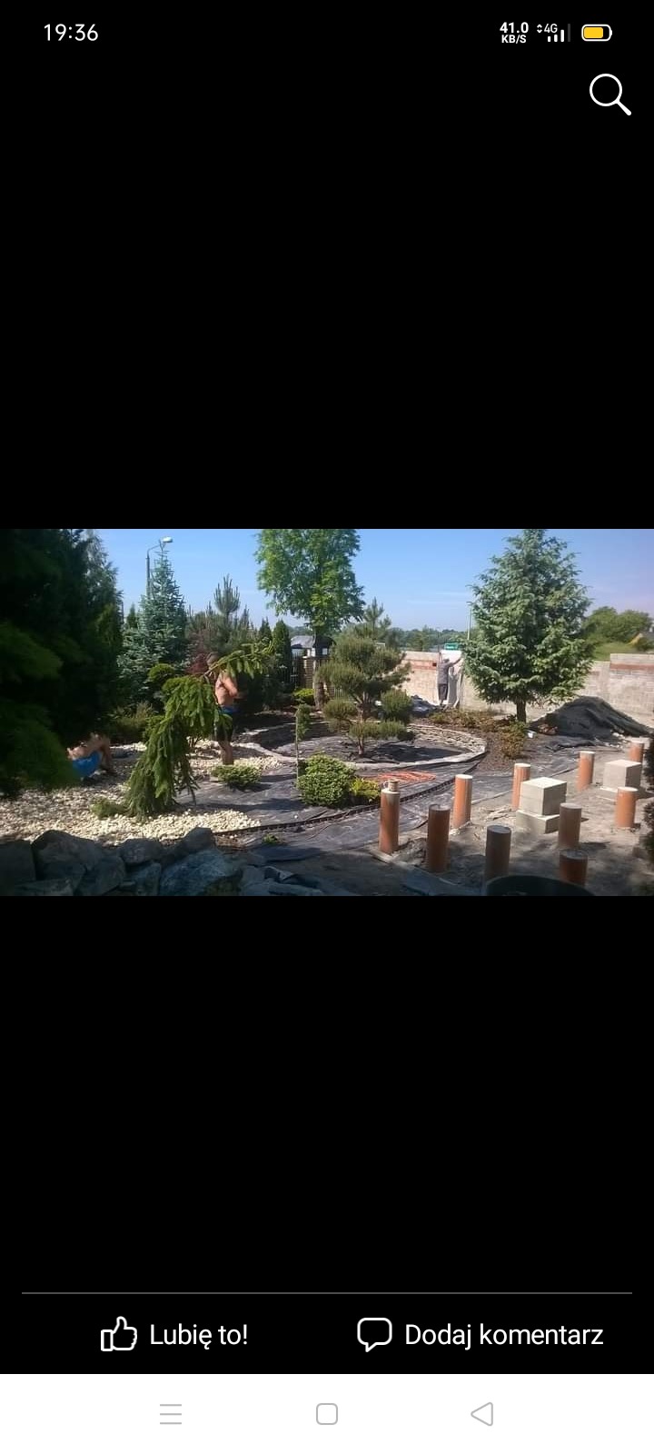 Prace ogrodnicze w toku: układanie kamieni, sadzenie drzew i krzewów iglastych, widoczne rury do systemu nawadniania.