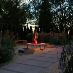 Ogród z paleniskiem otoczonym ozdobnymi trawami, kamienną ścieżką i minimalistyczną ławką.