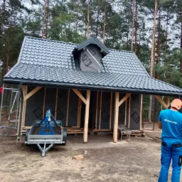 Niedokończony budynek z szarą blachodachówką, widoczna więźba dachowa i folia paroizolacyjna, przyczepka i pracownik w niebieskim stroju roboczym.