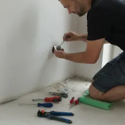 Elektryk klęczy przy ścianie, montując gniazdko elektryczne; widoczne narzędzia, fragment ściany z otworem na gniazdko i instalacja w trakcie podłączania.