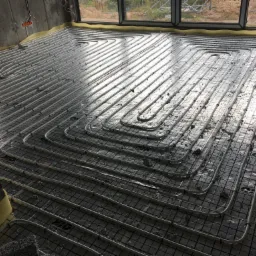 Rozłożone rury grzewcze podłogowe na izolacji z siatką metalową, widok z góry, surowy beton na ścianach i żółta taśma brzegowa.