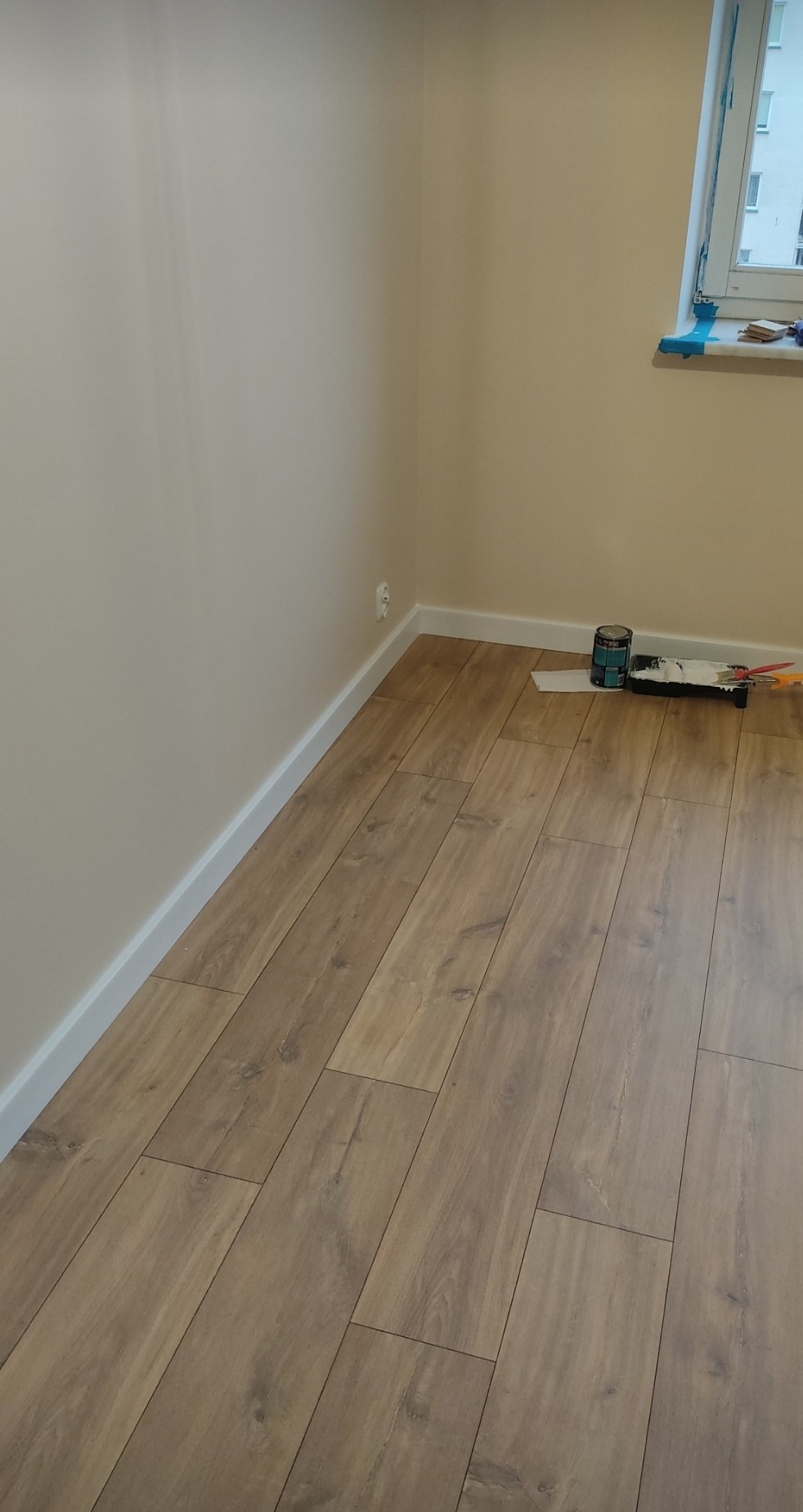Jasne panele podłogowe ułożone w pokoju, widoczne pomalowane ściany i listwy przypodłogowe oraz puszka z farbą i wałek malarski przy oknie.