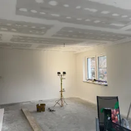 Wnętrze pokoju w trakcie remontu: sufit z zamontowaną płytą kartonowo-gipsową przed szpachlowaniem, ściany przygotowane do malowania, na podłodze widoczny bałagan remontowy, drabina i reflektor...